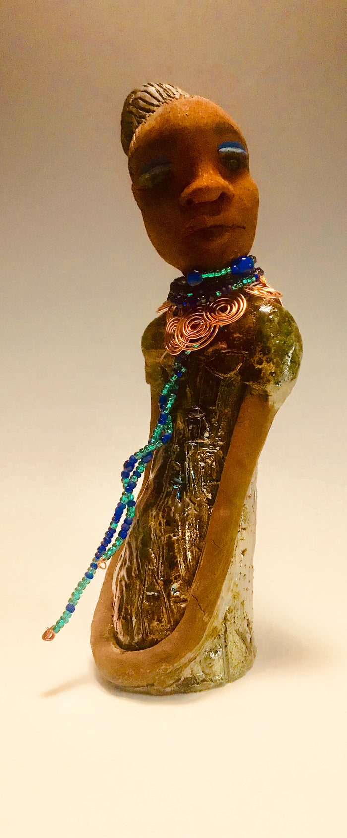 Emma stands 12.5" x3. 5" x 4" and weighs 2.04 lbs. She has a lovely honey brown complexion with  reddish brown lips. She has a braided hairstyle.  Emma has a colorful metallic green antique copper glazed dress. She wears a spiral copper wire necklace on top of a multicolored blue beaded collar. With eyes wide opened,  Emma has hopes of finding a new home.