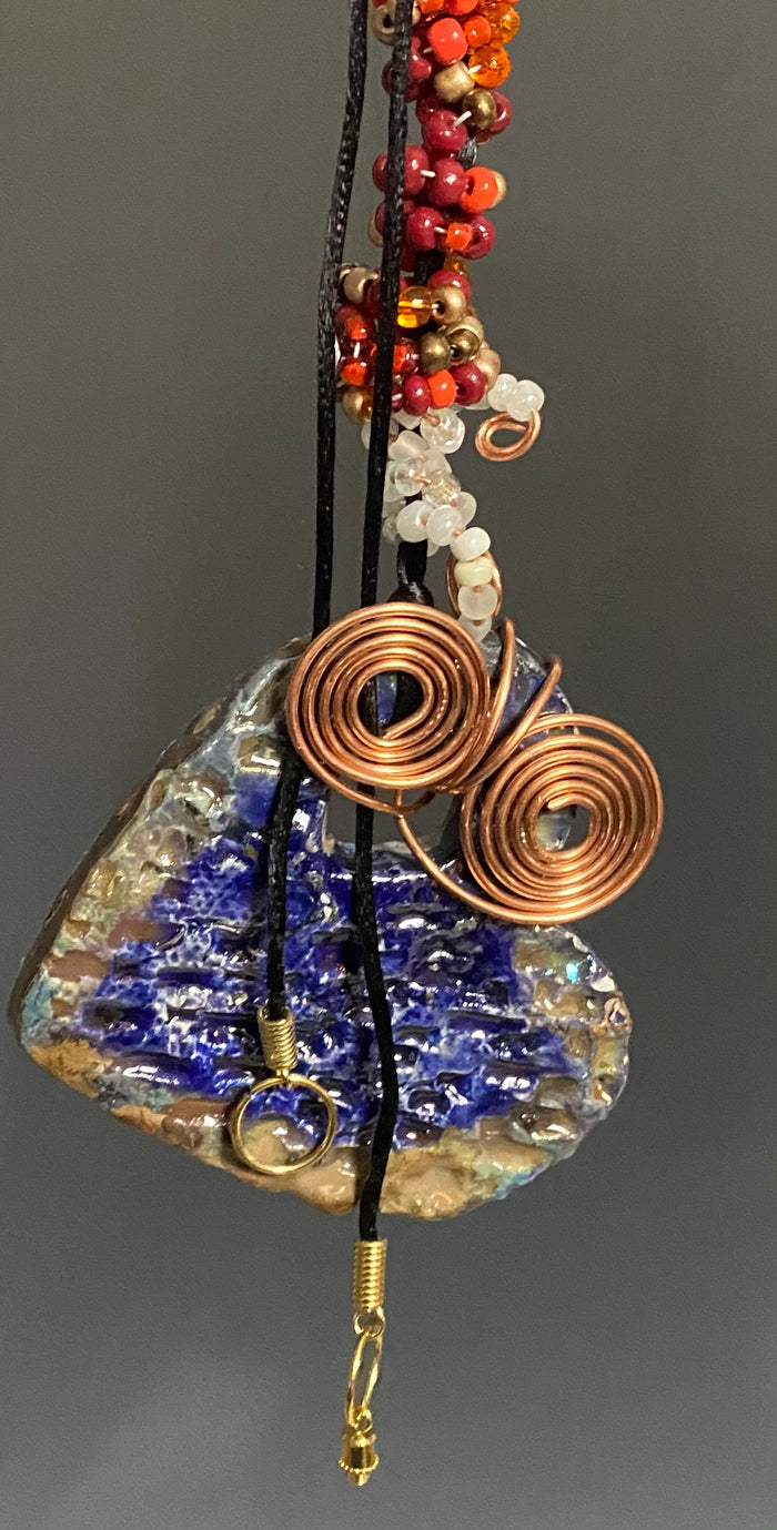 Have A Heart! Each heart pendant is handmade with love! It is 3"x 3"and weighs approx. 3ozs. This pendant has a royal blue and gold metallic raku glazes that renders a unique translucent  patina. The heart has a textured pattern . Both sides are  are different and equally beautiful! It holds a spiral of red  and white mini beads on a spiral copper wire. This pendant has a nice 12" black adjustable rattail cord!